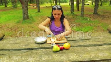 一位坐在一张木桌旁的公园里的年轻女子<strong>将一个</strong>苹果切成薄片作为水果沙拉。